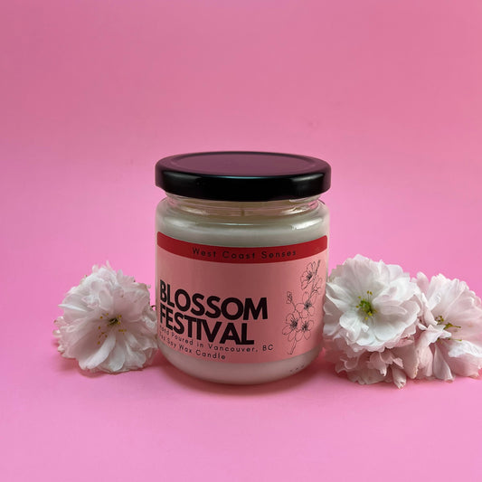Soy wax candle in a glass jar with a black lid. The label is pink and reads Blossom Festival. There are cherry blossoms on either side of the candle. The background is pink