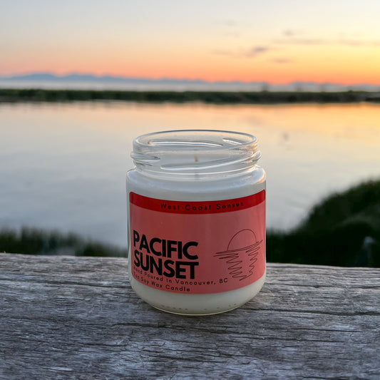 Soy wax candle in a glass jar with a red label that reads Pacific Sunset. The candle is on a log with an ocean view and an orange sky in the background
