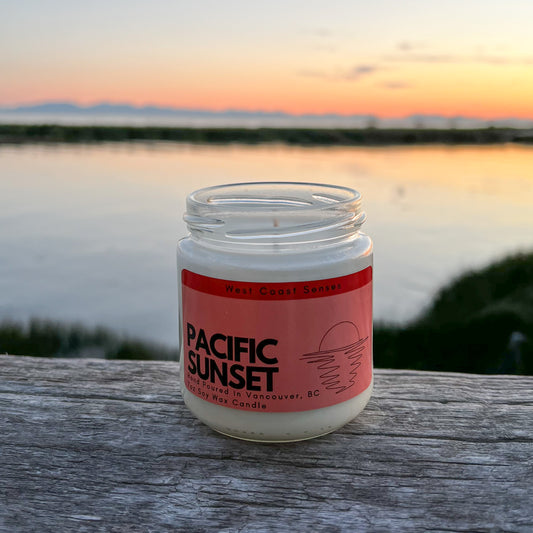 Soy wax candle in a glass jar with a red label that reads Pacific Sunset. The candle is on a log with an ocean view and an orange sky in the background